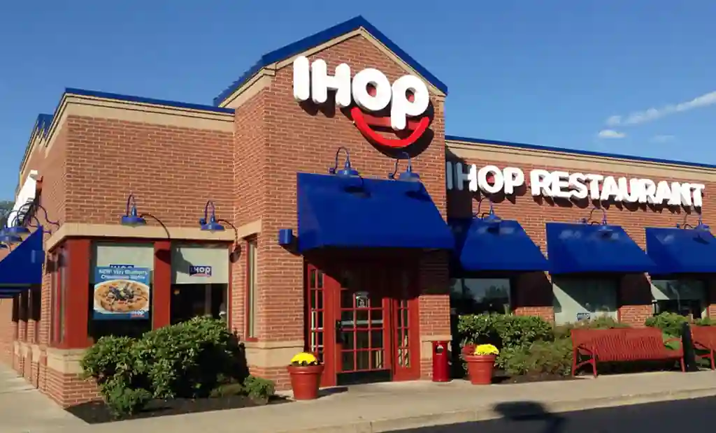 Front view of an IHOP restaurant with red brick walls, blue awnings, and signage, including a poster advertising blueberry cheesecake waffles.