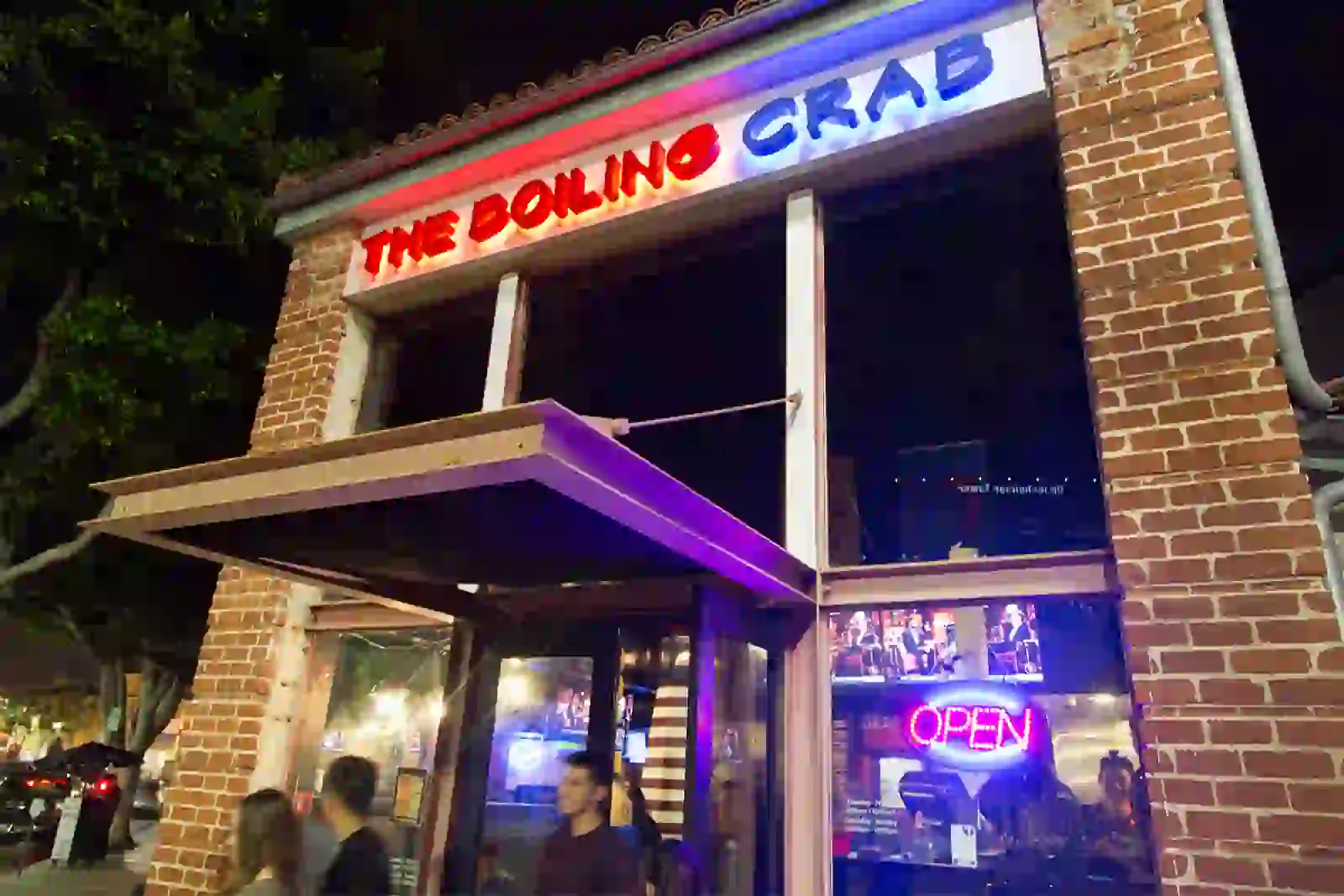 Exterior of The Boiling Crab restaurant at night, featuring a brick facade, neon signs, and large windows showing a lively dining scene inside.