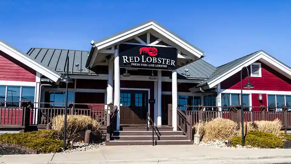 Front exterior of Red Lobster restaurant with red and white facade, signature signage featuring a red lobster logo, the tagline “FRESH FISH • LIVE LOBSTER,” and landscaped entryway with stairs.