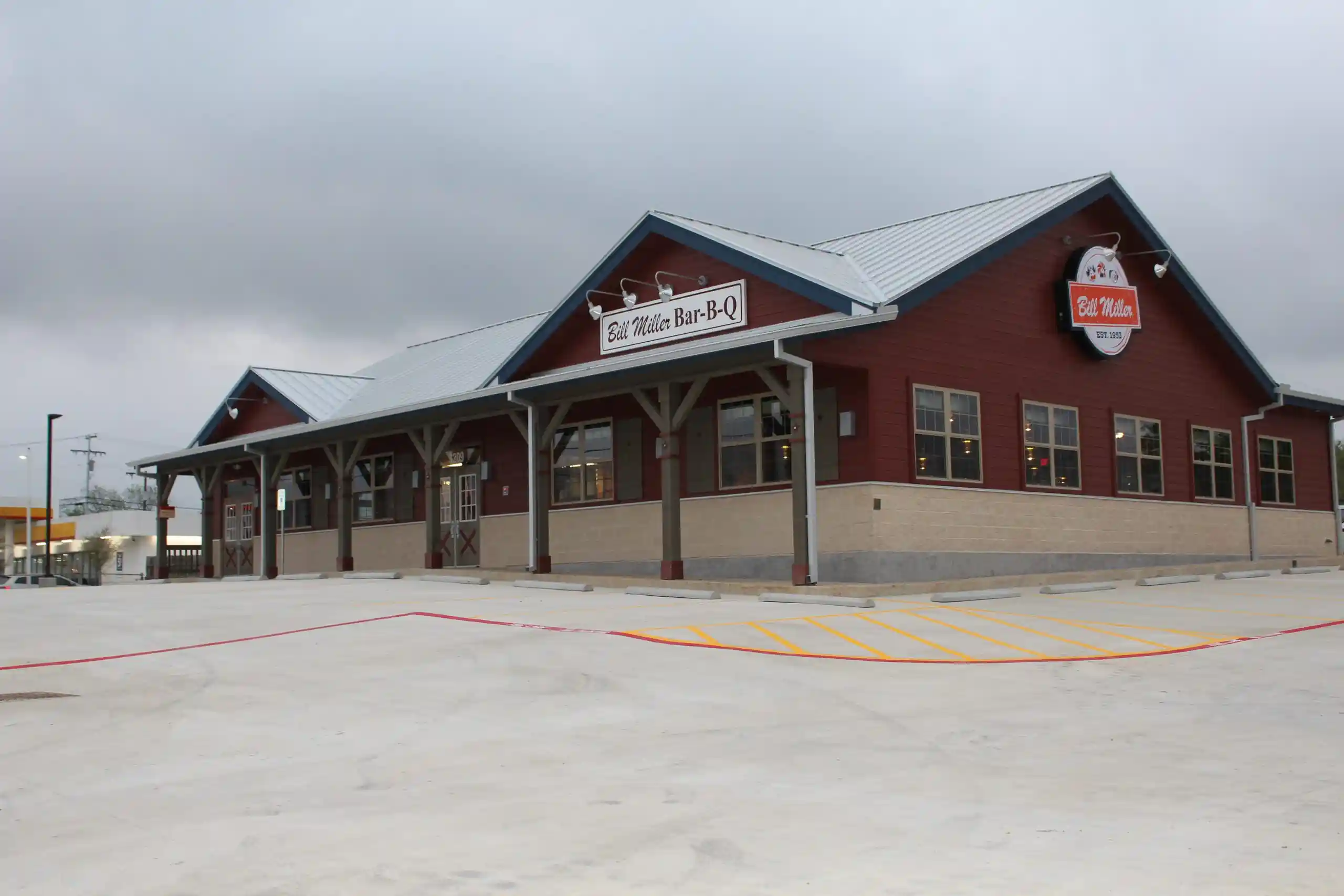 Exterior view of Bill Miller Bar-B-Q restaurant with brick and wood design, pitched roof, and covered walkway in front of empty parking lot under cloudy skies.