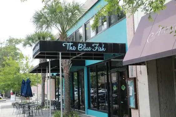 Exterior of The Blue Fish restaurant with teal walls, black awning, cursive signage, palm trees, and outdoor seating along a city sidewalk.