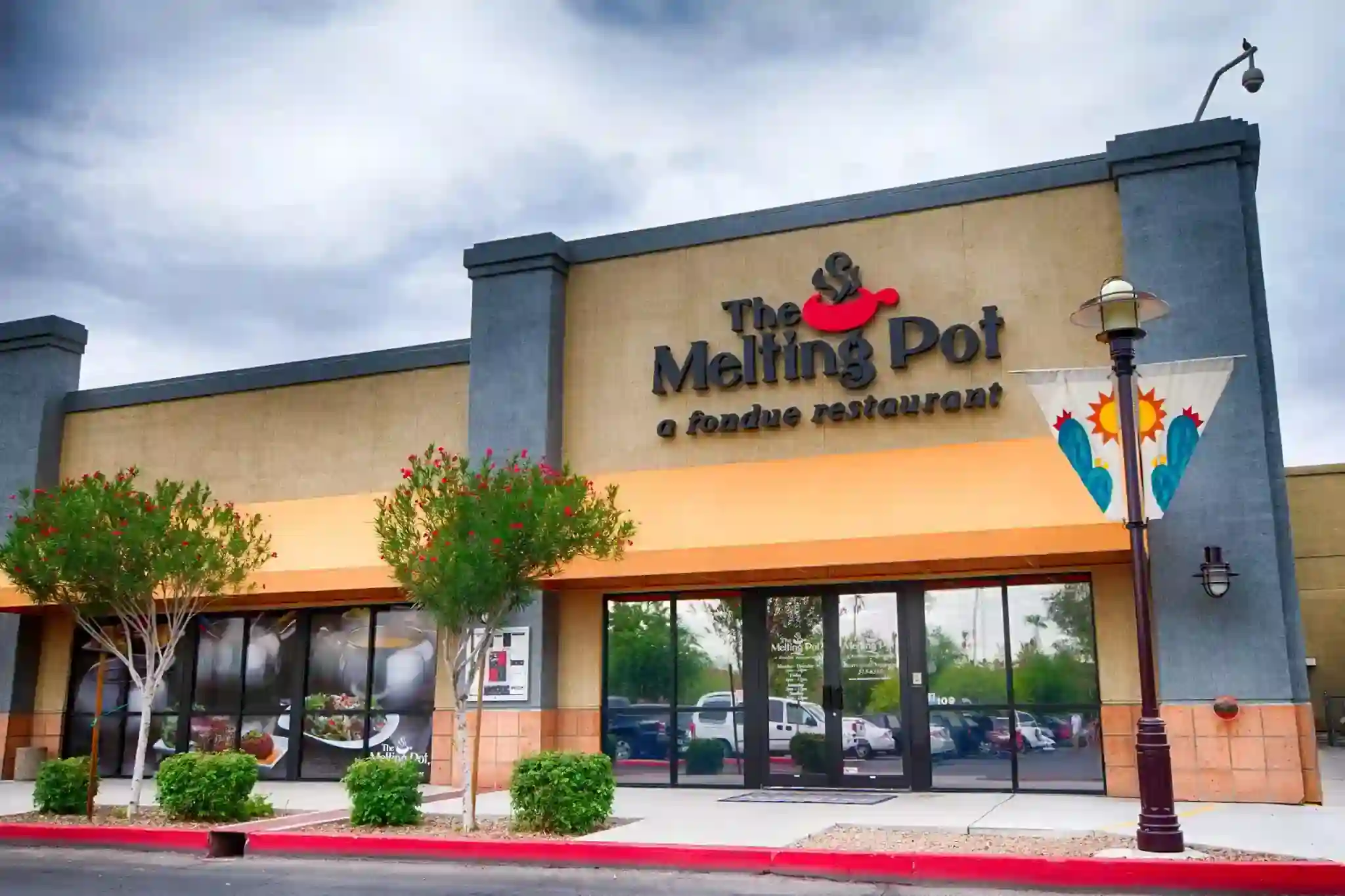 The Melting Pot fondue restaurant exterior with beige and gray facade, orange awning, red floral landscaping, and a decorative streetlamp banner featuring a sun and cacti.