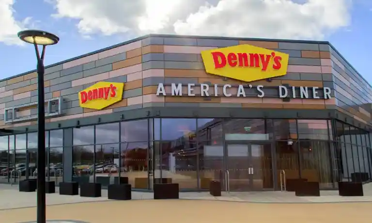 Exterior of a modern Denny’s restaurant featuring beige and gray panels, red and yellow Denny’s signage, large glass windows, and “AMERICA’S DINER” text above the entrance.