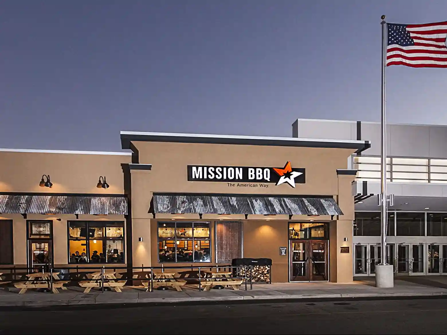 Evening view of MISSION BBQ restaurant with lighted signage, American flag, outdoor picnic tables, and corrugated metal awnings under a dark sky.