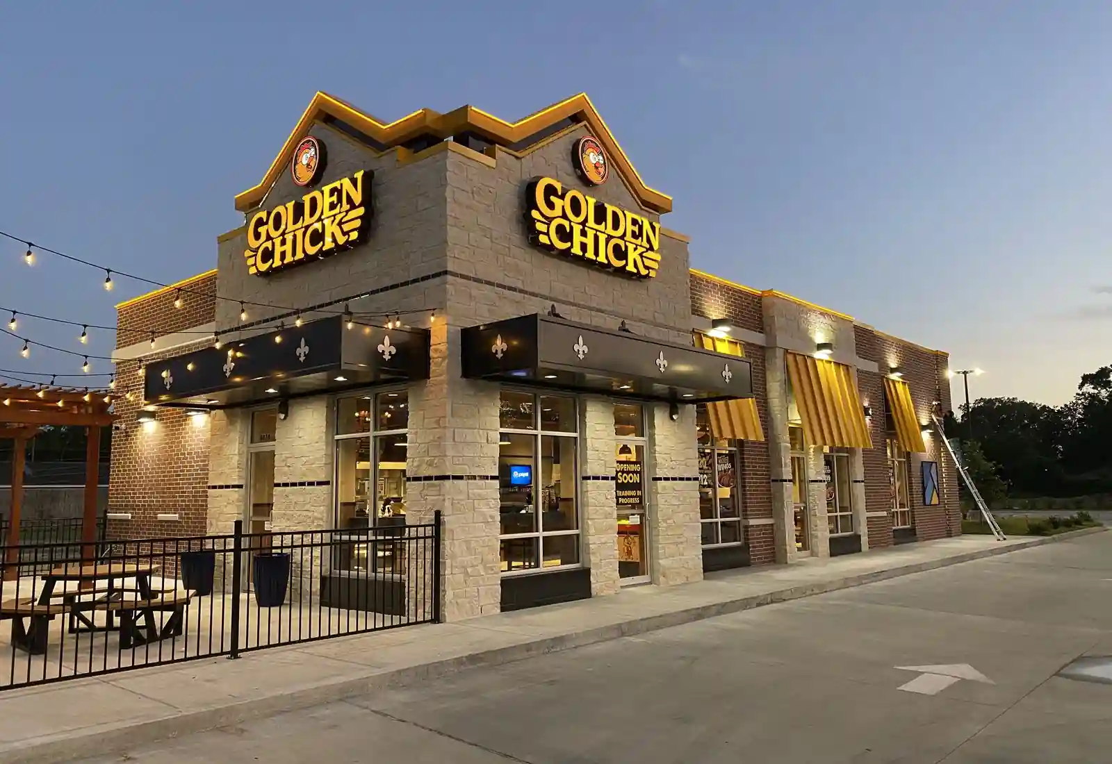 Exterior of a Golden Chick restaurant at dusk, with brick and stone facade, illuminated signage, patio picnic tables, string lights, and a sign on the door reading "Opening Soon Training in Progress.