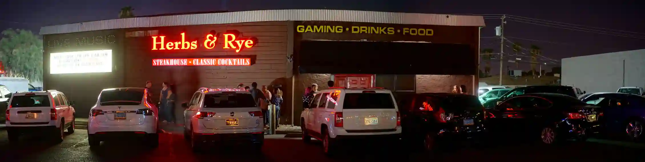 A nighttime view of the exterior of Herbs & Rye steakhouse and cocktail bar, with illuminated signage, parked cars, and patrons standing near the entrance.