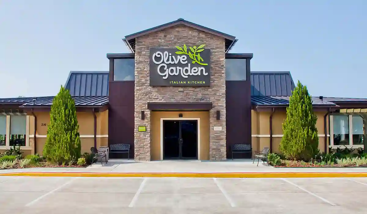 Olive Garden Italian Kitchen restaurant exterior with stone accent facade, white “Olive Garden” signage with green olive branch logo, landscaped entryway, and benches near the entrance under an overcast sky.