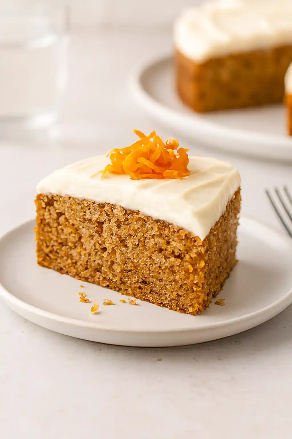 Moist gluten free carrot cake slice topped with cream cheese frosting and carrot shavings, plated in bright natural kitchen lighting.